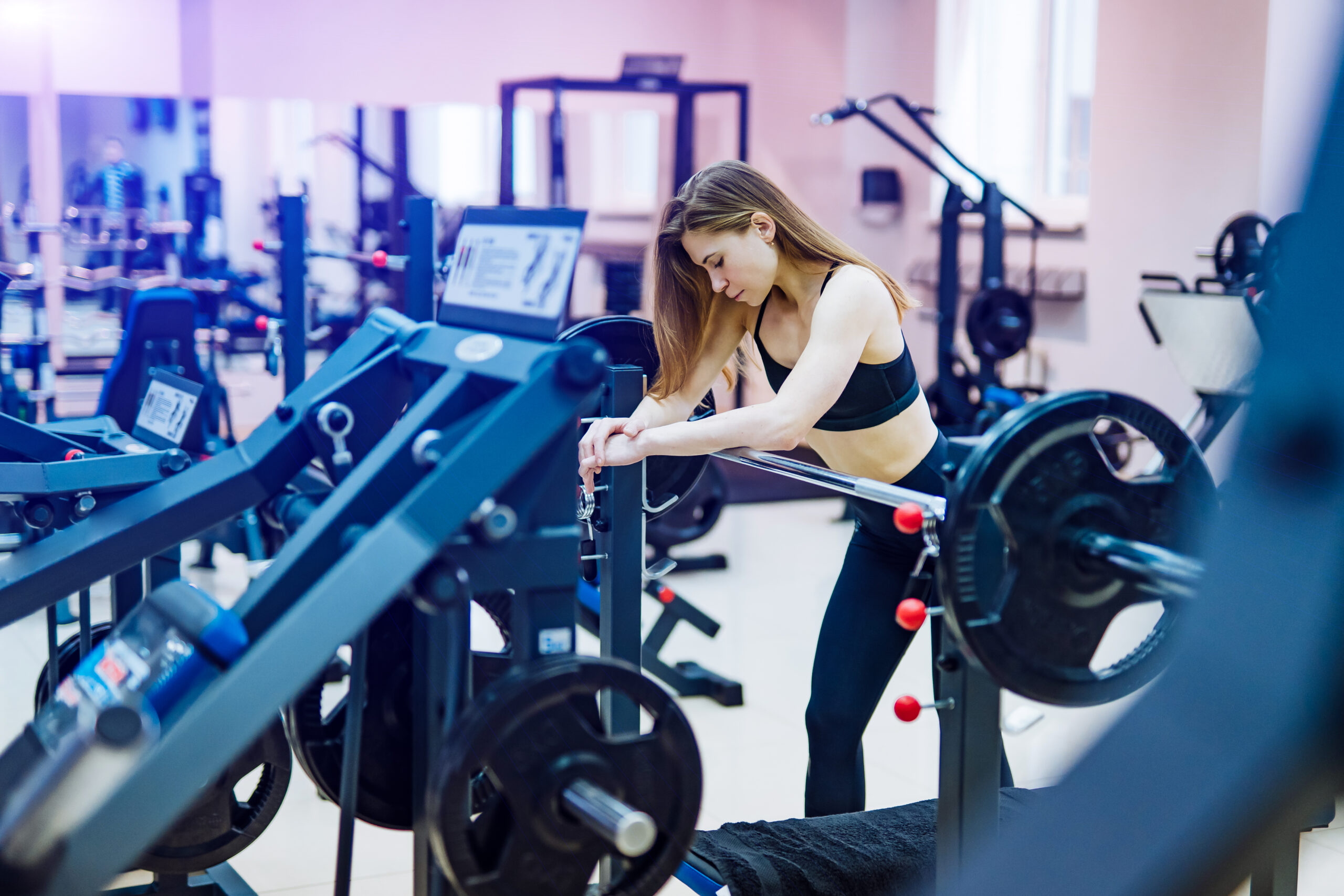 Beautiful muscular fitness woman in a black sporty clothes leaning on simulator with barbell at gym. Athletic girl resting after sport workout exercises among sports equipment